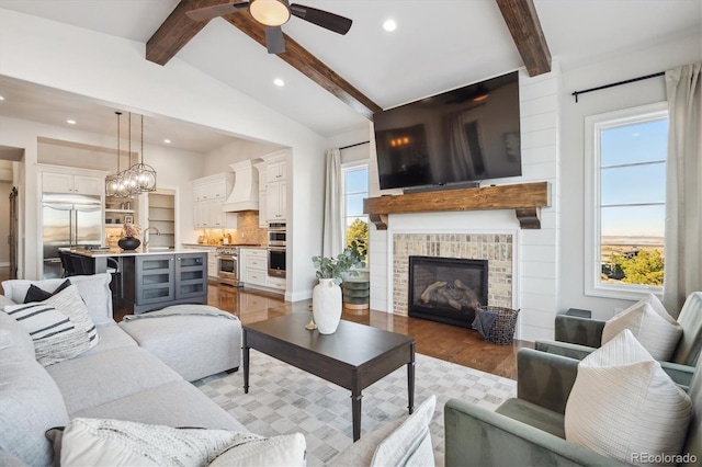 living room with a brick fireplace, ceiling fan with notable chandelier, sink, lofted ceiling with beams, and light hardwood / wood-style floors