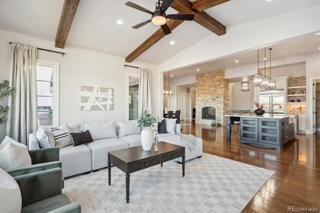 living room featuring light wood-type flooring, ceiling fan with notable chandelier, sink, vaulted ceiling with beams, and a stone fireplace