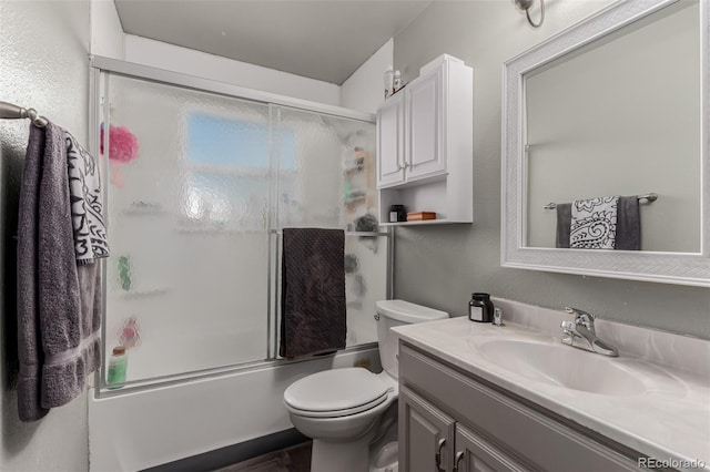 full bath featuring toilet, a textured wall, bath / shower combo with glass door, and vanity