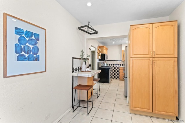 kitchen with light tile patterned flooring, light brown cabinetry, tile countertops, appliances with stainless steel finishes, and a kitchen breakfast bar