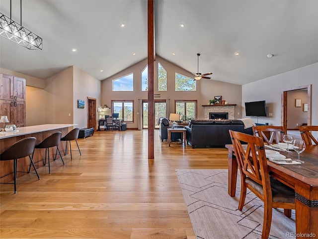 dining space featuring beamed ceiling, high vaulted ceiling, a ceiling fan, a lit fireplace, and light wood finished floors