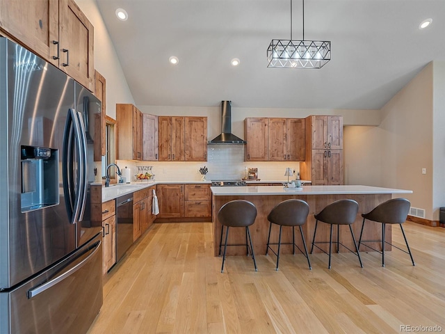 kitchen featuring wall chimney range hood, stainless steel appliances, brown cabinetry, light wood finished floors, and decorative backsplash