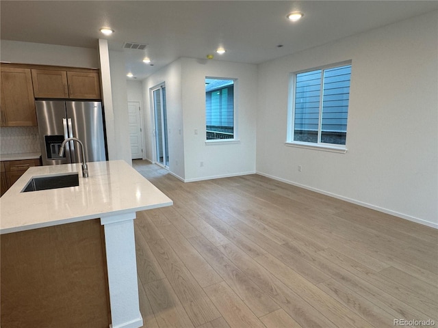 kitchen featuring stainless steel fridge, light wood-type flooring, backsplash, sink, and a center island with sink