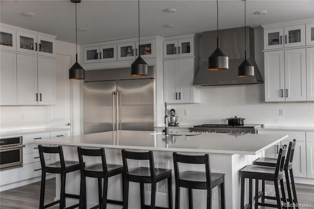 kitchen featuring wall chimney range hood, stainless steel appliances, an island with sink, decorative backsplash, and decorative light fixtures