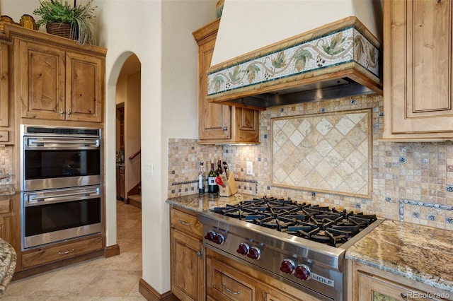 kitchen featuring light tile patterned floors, appliances with stainless steel finishes, light stone counters, decorative backsplash, and custom exhaust hood