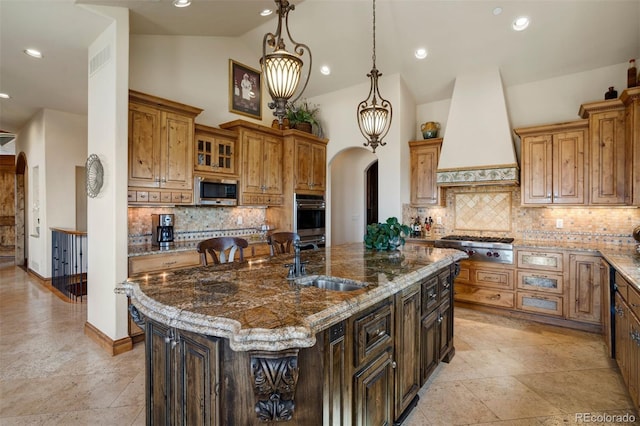 kitchen with decorative light fixtures, an island with sink, sink, stainless steel appliances, and custom range hood