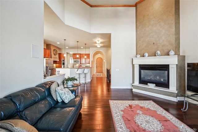 living room with dark wood-style flooring, a glass covered fireplace, baseboards, a towering ceiling, and ornamental molding