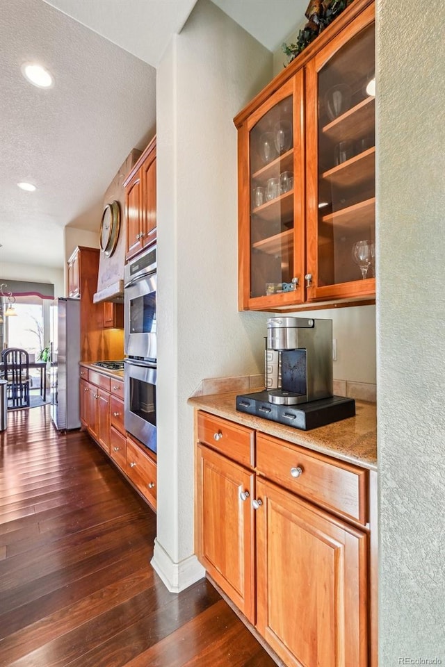 kitchen featuring appliances with stainless steel finishes, dark wood-style flooring, glass insert cabinets, a textured ceiling, and brown cabinetry