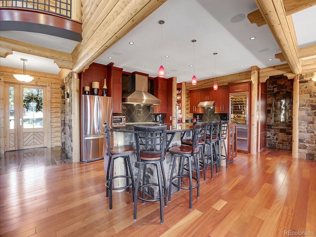 kitchen with wall chimney exhaust hood, decorative light fixtures, stainless steel fridge, log walls, and light hardwood / wood-style floors