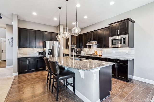 kitchen with under cabinet range hood, stainless steel appliances, wood finished floors, a sink, and light stone countertops