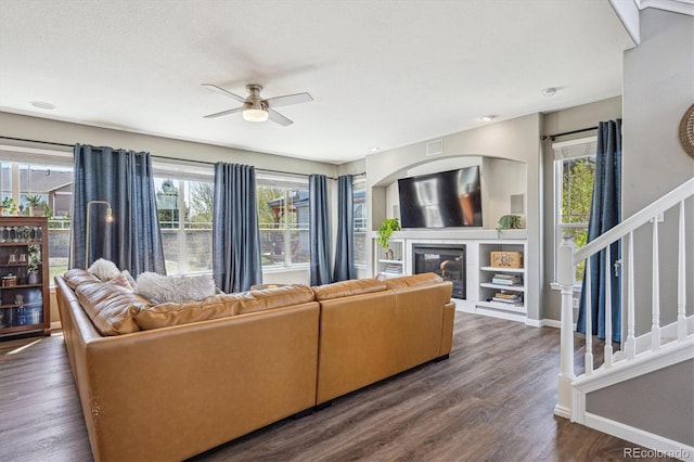 living area with dark wood-style flooring, a glass covered fireplace, stairs, baseboards, and a ceiling fan