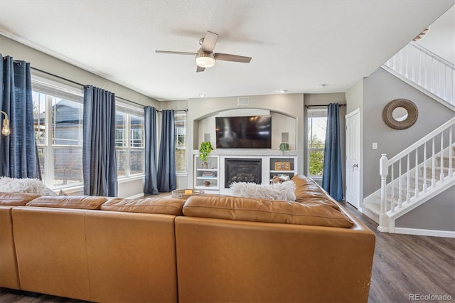 living room with stairs, a glass covered fireplace, wood finished floors, baseboards, and a ceiling fan