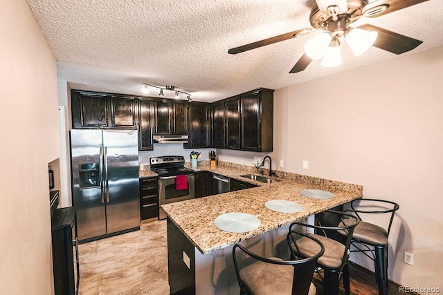 kitchen with light stone counters, under cabinet range hood, stainless steel appliances, a peninsula, and a sink