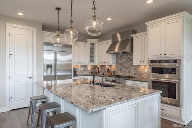 kitchen featuring stainless steel appliances, wall chimney range hood, a sink, decorative backsplash, and light stone counters