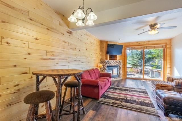 living room with wood walls, wood-type flooring, ceiling fan with notable chandelier, and a stone fireplace