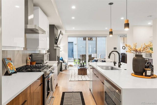 kitchen with hanging light fixtures, stainless steel appliances, light hardwood / wood-style flooring, sink, and wall chimney exhaust hood