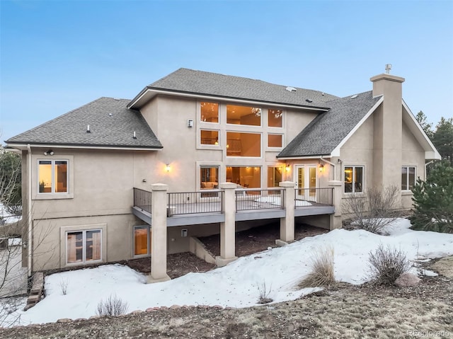 snow covered rear of property featuring a deck, roof with shingles, a chimney, and stucco siding