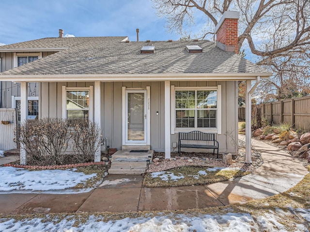 snow covered property entrance with board and batten siding, roof with shingles, fence, and a porch