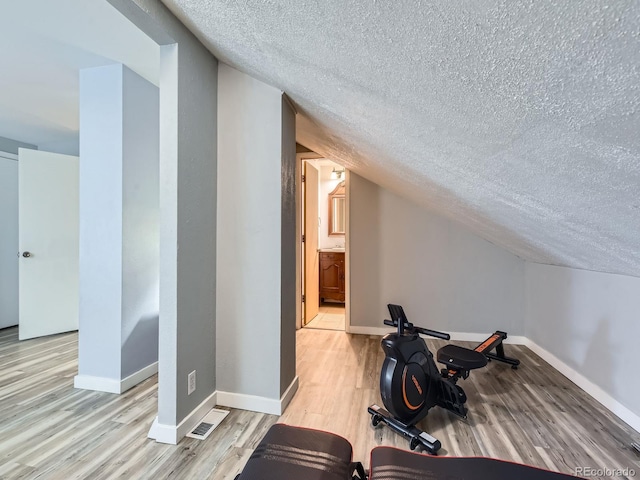 workout room featuring light wood finished floors, baseboards, vaulted ceiling, and a textured ceiling