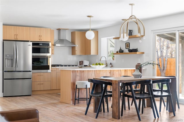 kitchen featuring light wood finished floors, open shelves, stainless steel appliances, light brown cabinets, and wall chimney exhaust hood