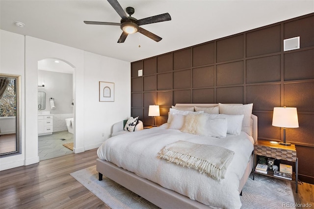 bedroom with wood finished floors, arched walkways, a decorative wall, baseboards, and a ceiling fan