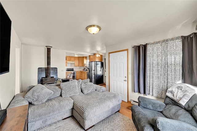 living room featuring a baseboard heating unit, a wood stove, light wood finished floors, and ceiling fan