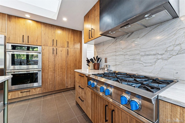 kitchen featuring dark tile patterned flooring, backsplash, stainless steel appliances, a skylight, and wall chimney range hood