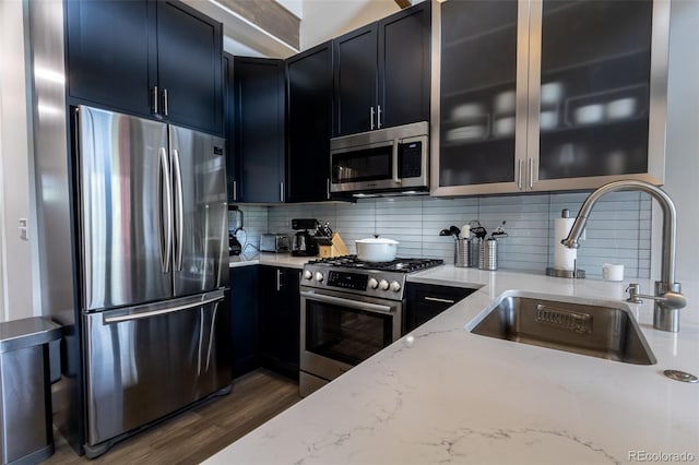 kitchen with a sink, dark cabinetry, appliances with stainless steel finishes, light stone countertops, and glass insert cabinets