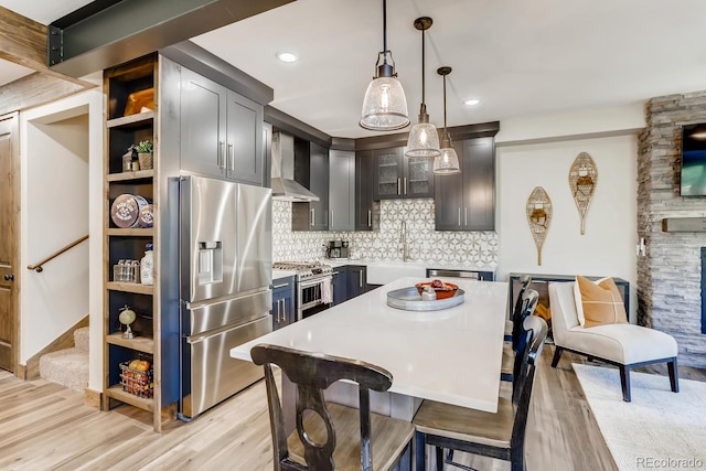 kitchen with stainless steel appliances, light countertops, wall chimney exhaust hood, light wood finished floors, and decorative light fixtures