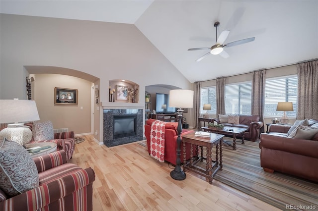living area with a ceiling fan, arched walkways, high vaulted ceiling, light wood-style floors, and a premium fireplace
