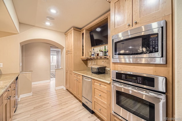 kitchen with arched walkways, appliances with stainless steel finishes, light stone countertops, light brown cabinetry, and light wood-style floors