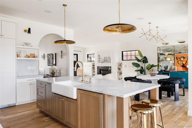 kitchen with a sink, light wood-type flooring, open shelves, an island with sink, and decorative light fixtures