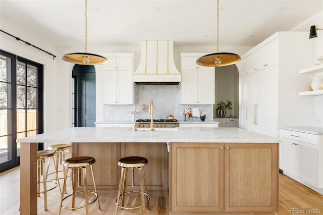 kitchen with light wood finished floors, custom exhaust hood, tasteful backsplash, light stone countertops, and a large island with sink