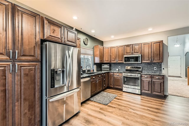 kitchen featuring appliances with stainless steel finishes, backsplash, dark stone countertops, light wood-type flooring, and a sink