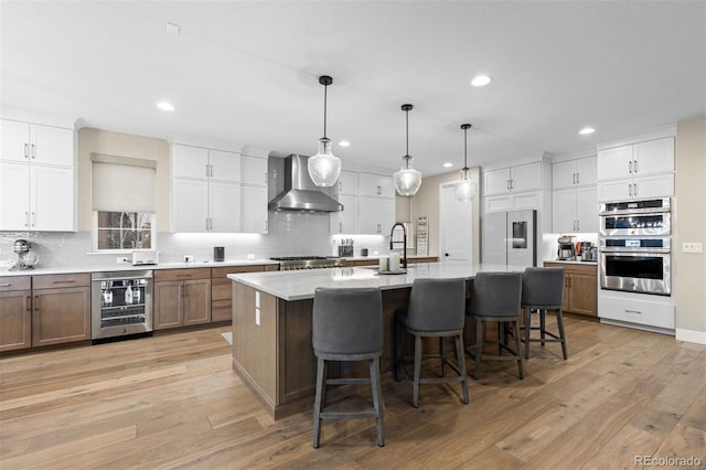 kitchen with wine cooler, appliances with stainless steel finishes, a large island with sink, wall chimney range hood, and decorative light fixtures