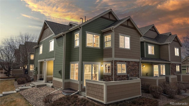 back of house at dusk with stone siding and a shingled roof