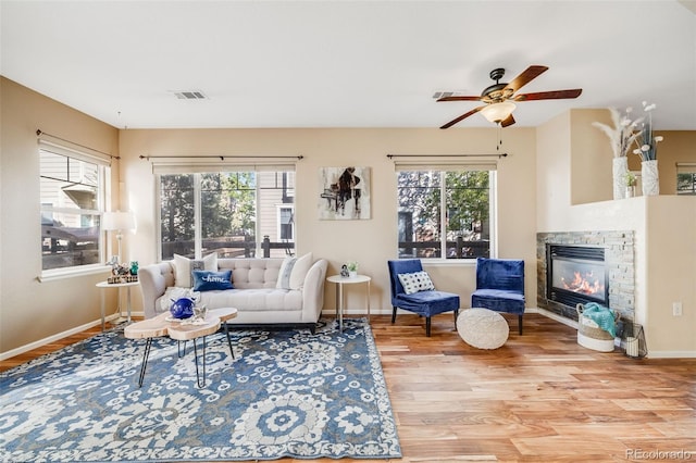 living area featuring wood finished floors, a stone fireplace, a ceiling fan, and baseboards