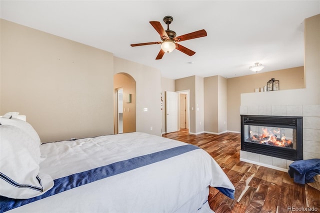 bedroom with wood finished floors, arched walkways, a fireplace, and ceiling fan