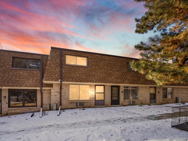 snow covered property featuring roof with shingles, brick siding, and mansard roof