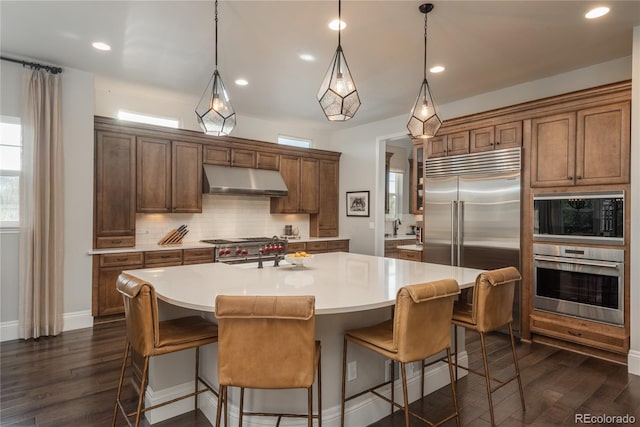 kitchen featuring decorative backsplash, built in appliances, hanging light fixtures, and a center island with sink