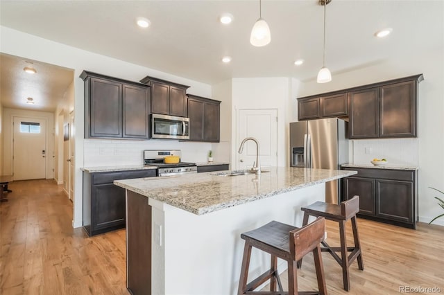 kitchen featuring sink, appliances with stainless steel finishes, a kitchen island with sink, hanging light fixtures, and light wood-type flooring