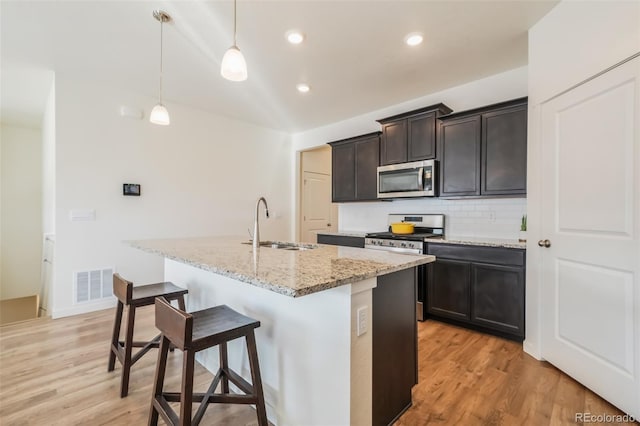 kitchen with sink, decorative light fixtures, appliances with stainless steel finishes, light stone countertops, and a kitchen island with sink