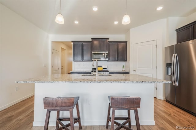 kitchen featuring stainless steel appliances, an island with sink, hanging light fixtures, and light stone counters