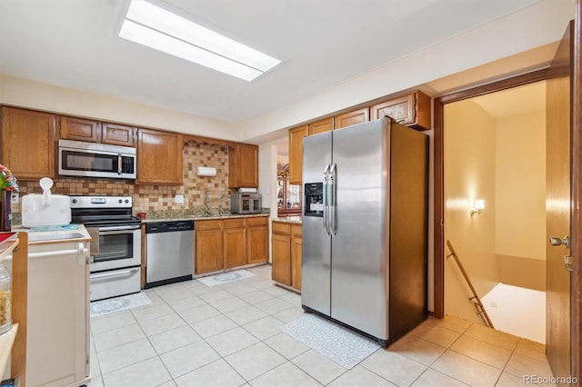 kitchen featuring light countertops, backsplash, appliances with stainless steel finishes, brown cabinetry, and light tile patterned flooring