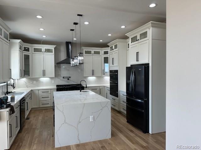 kitchen featuring a center island with sink, a sink, black appliances, wood finished floors, and wall chimney exhaust hood