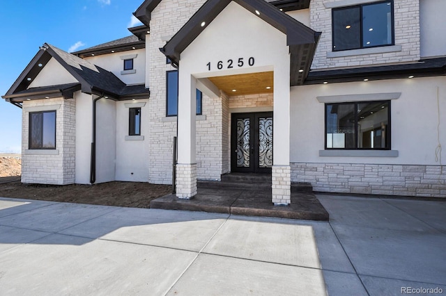 view of exterior entry featuring stone siding, stucco siding, french doors, and a shingled roof