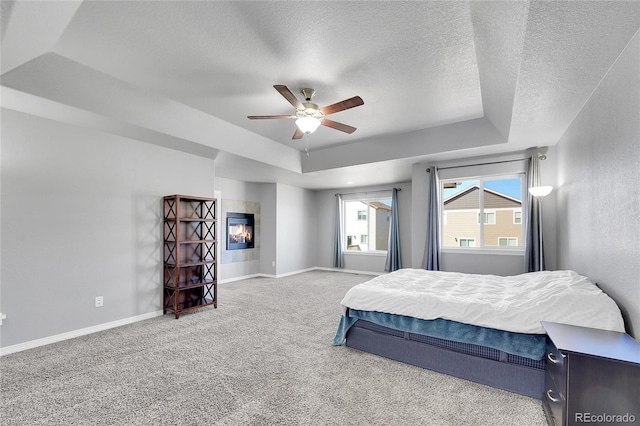 carpeted bedroom with baseboards, a ceiling fan, a textured ceiling, a tray ceiling, and a glass covered fireplace