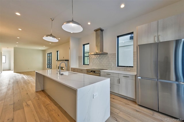 kitchen featuring wall chimney exhaust hood, stainless steel appliances, decorative light fixtures, a kitchen island with sink, and light wood-type flooring