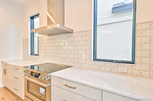 kitchen featuring high end range, light hardwood / wood-style flooring, wall chimney exhaust hood, light stone countertops, and tasteful backsplash