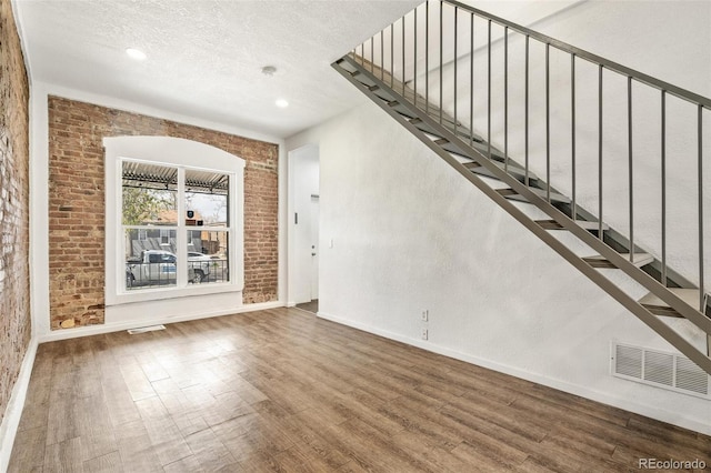 unfurnished living room featuring wood finished floors, baseboards, stairway, recessed lighting, and brick wall